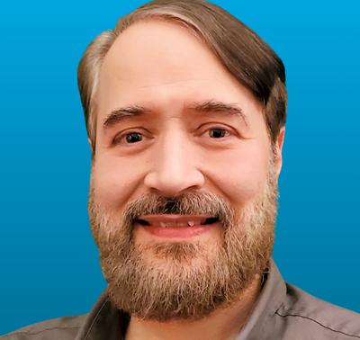 Portrait of Carl Burkart against a blue background, featuring a smiling man with a beard, showcasing professionalism and approachability.