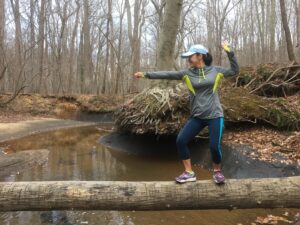Liani hiking, balancing on a log over a stream in the woods. New MMI Direct team member enjoying outdoor activities.